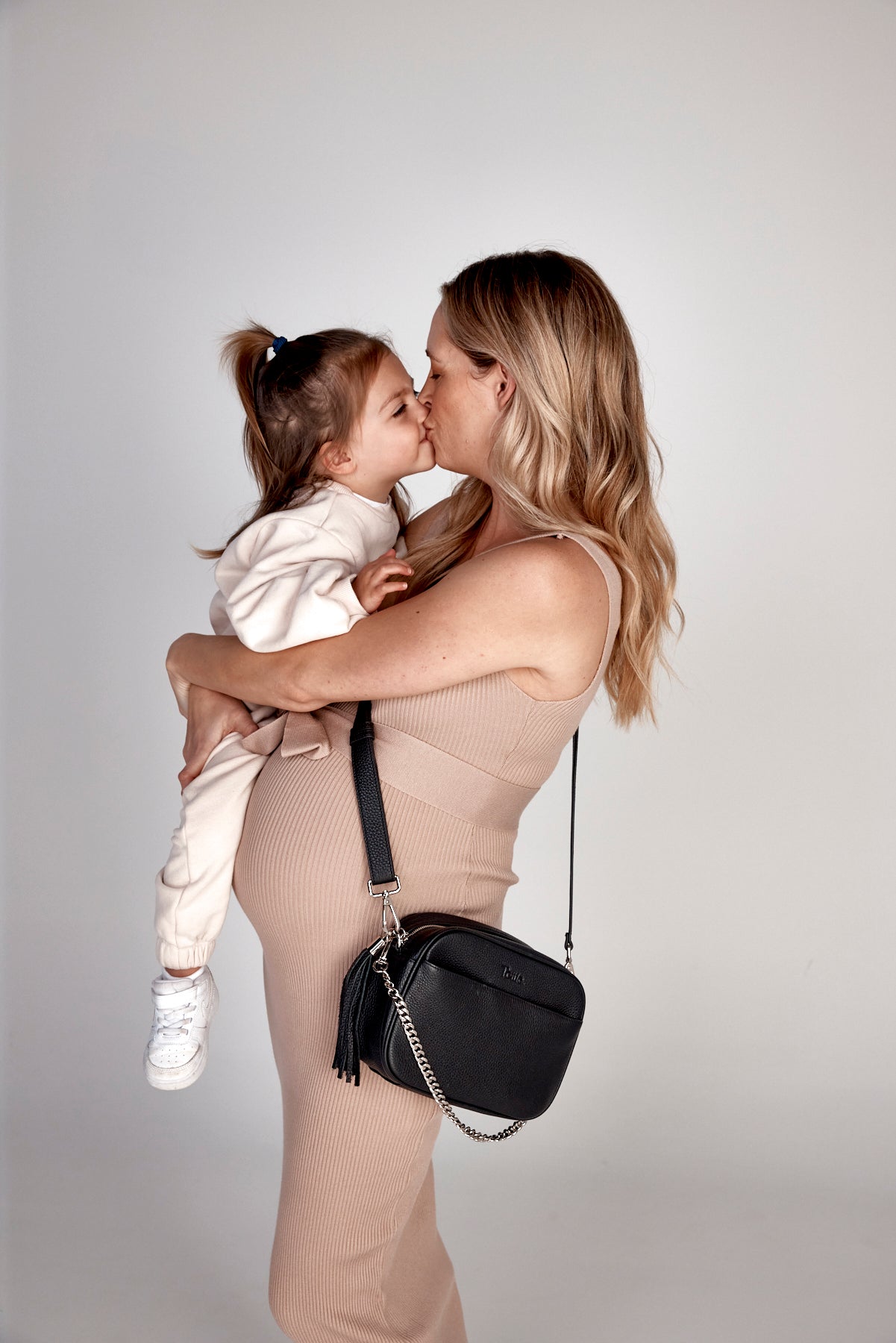 A pregnant woman kisses her toddler while wearing a black crossbody bag with a silver chain.