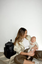 Woman sitting, carrying a baby and wearing a black nappy backpack from Tottie