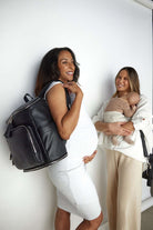 A pregnant woman carrying a black leather nappy backpack with silver hardware, standing alongside her friend who is carrying a baby and a black crossbody bag from Tottie