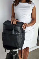 A woman in a white dress displaying a black leather nappy backpack with black zippers.