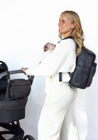 Woman carrying a baby and holding a black stroller. The stroller has a black crossbody bag from Tottie attached to the black pram clips. The woman is also wearing a black nappy backpack from Tottie