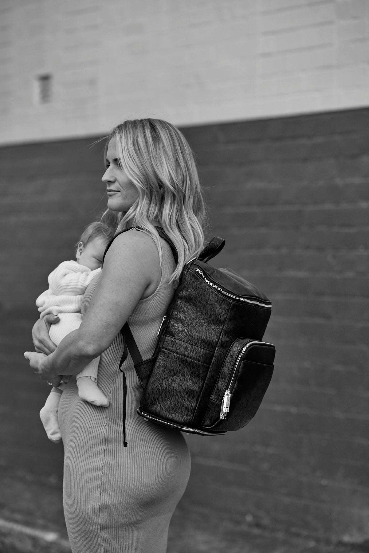 Black and white image of a woman holding a baby while carrying a black leather nappy backpack with silver hardware