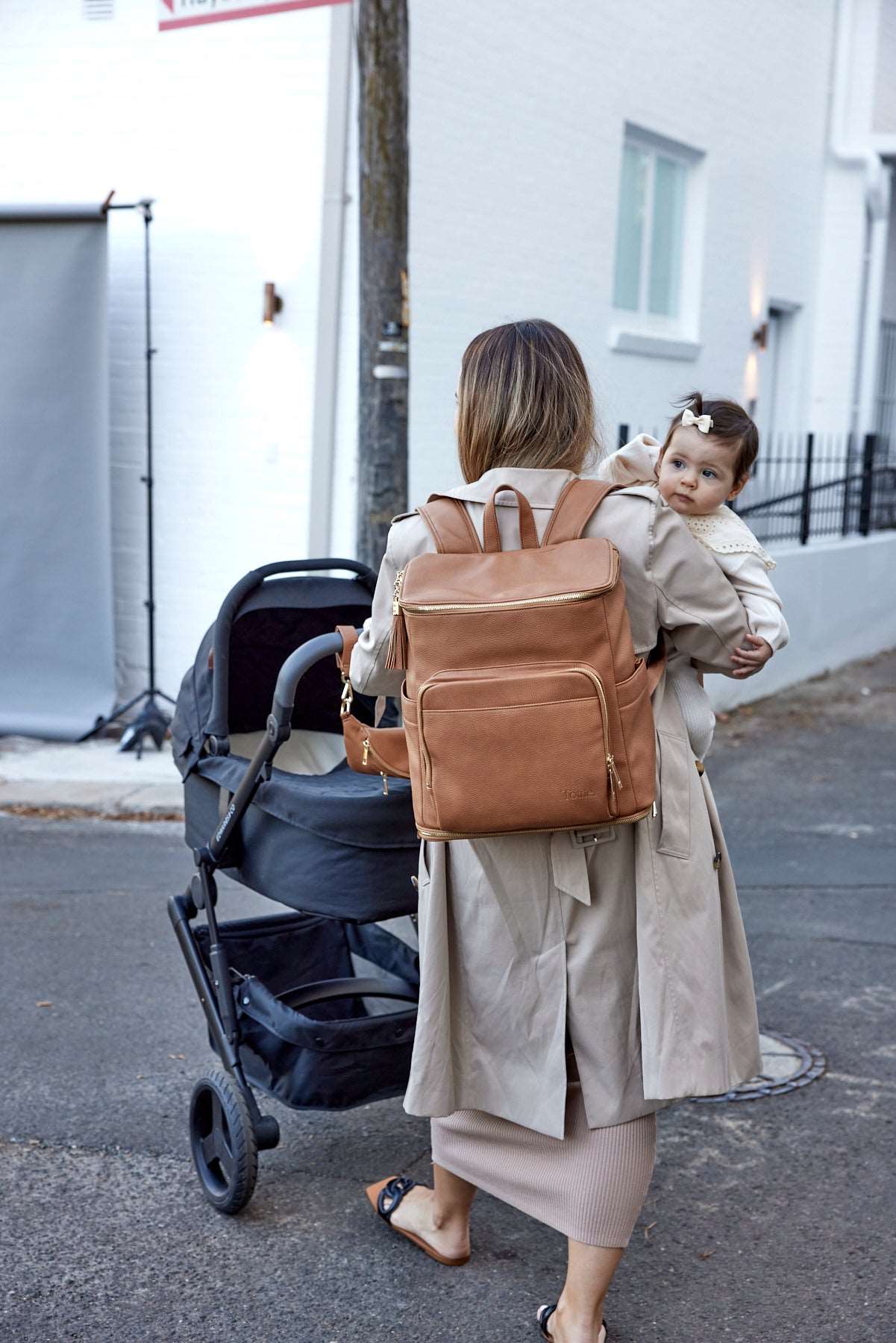 Woman carrying a baby and pushing a black stroller with Tottie's tan pram clips and bum bag. The woman is also wearing a tan nappy bag from Tottie.