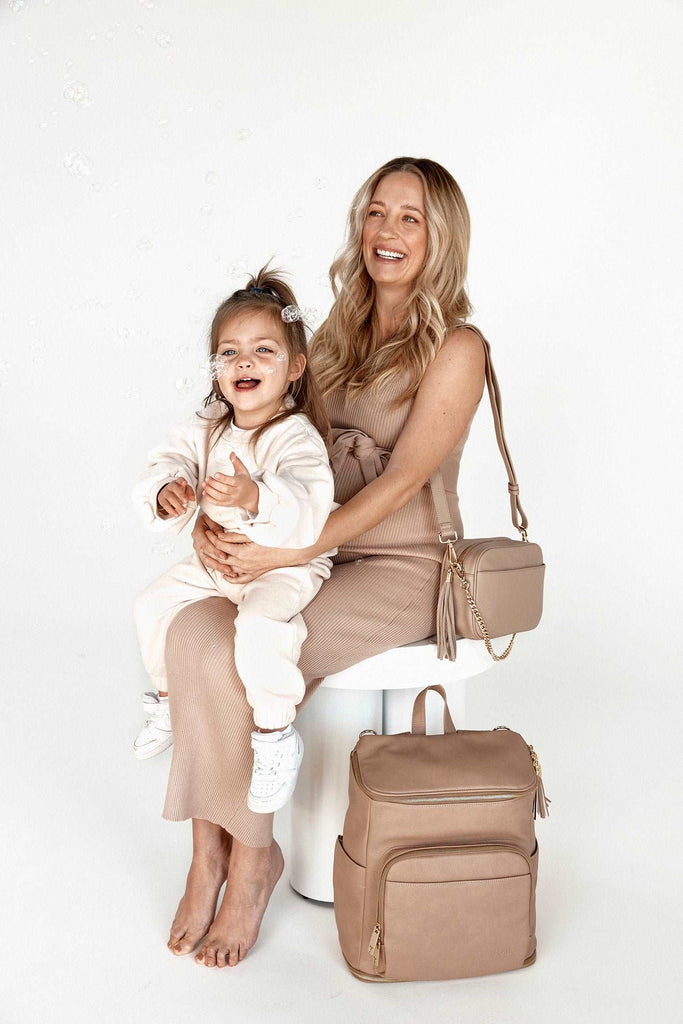 A woman and a child are seated on a stool next to a stone colour leather baby backpack