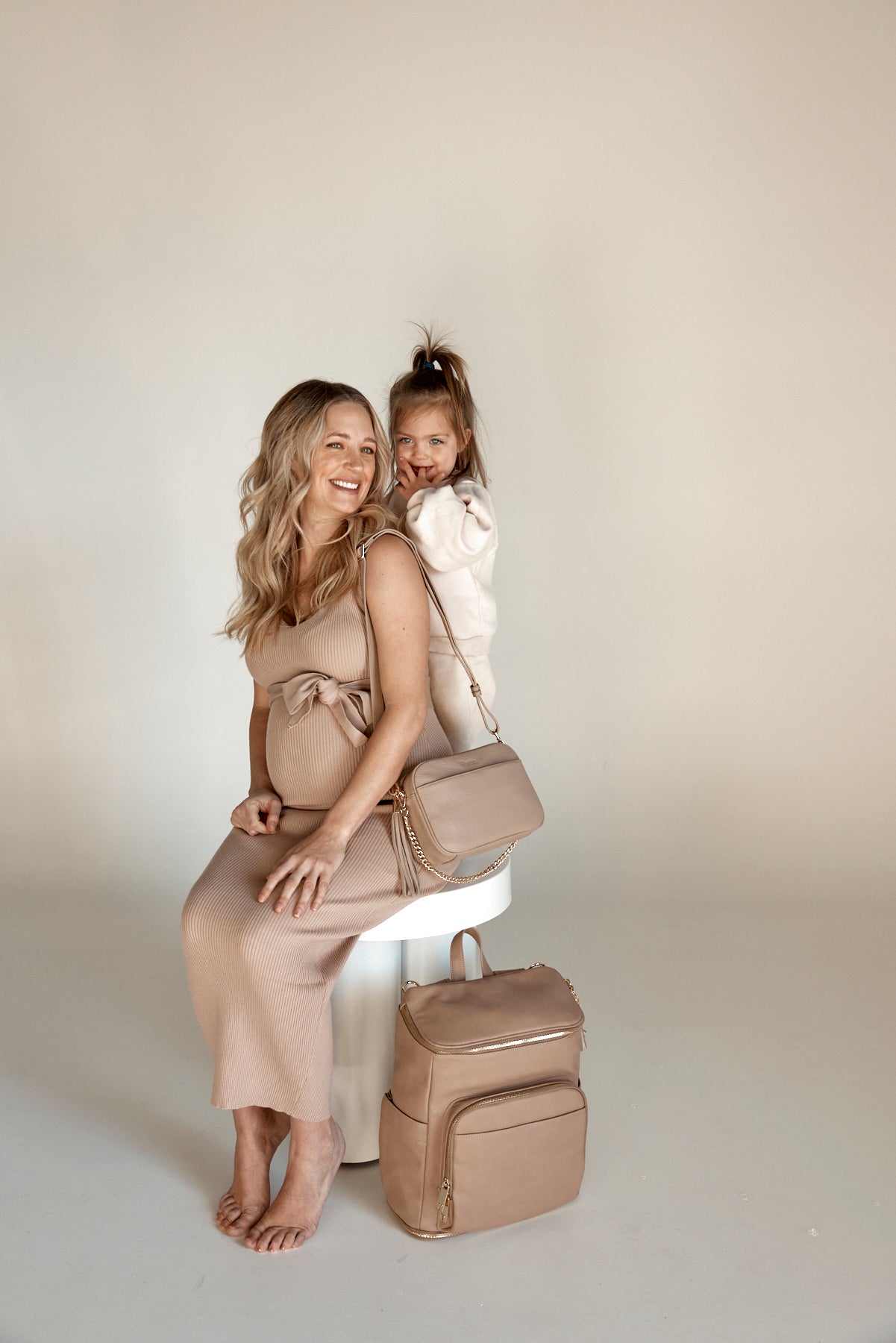 a pregnant woman and a child on her back while carrying a stone bestie crossbody bag and stone nappy bag backpack from Tottie