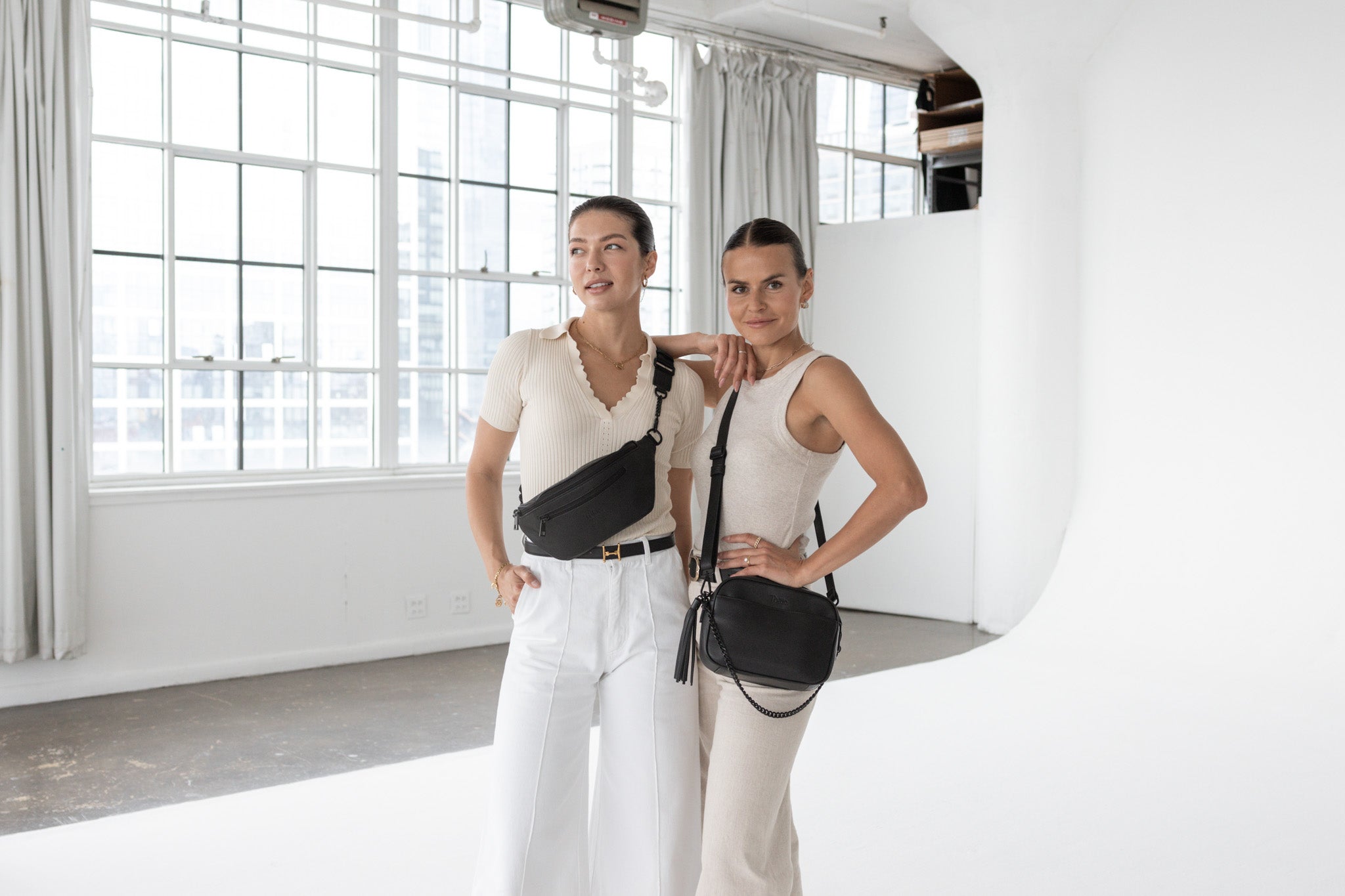 Two women wearing white outfits. One is wearing a black bum bag from Tottie, and the other is carrying a black crossbody bag from Tottie.