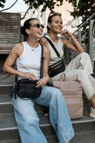 Two women sitting on stairs. One woman is wearing a black crossbody bag from Tottie, the other is wearing a black belt bag from Tottie, with Tottie's stone nappy backpack on the side