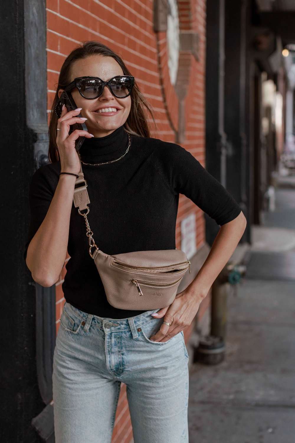 A woman in jeans and a black top standing on the street carrying a bum bag