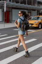 A woman with a black baby bag backpack crosses the street in a crosswalk