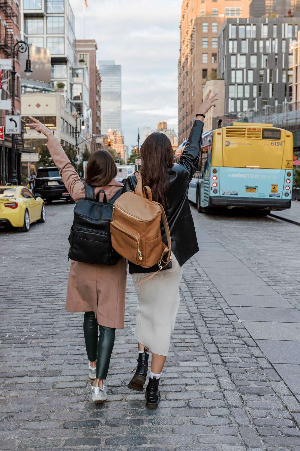 Two women stroll down a street, each carrying a leather baby backpack