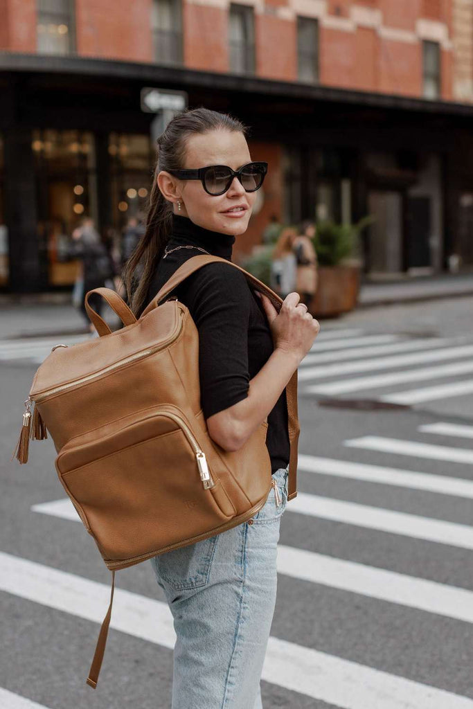  A woman in sunglasses carries a tan baby backpack, enjoying a sunny day outdoors.